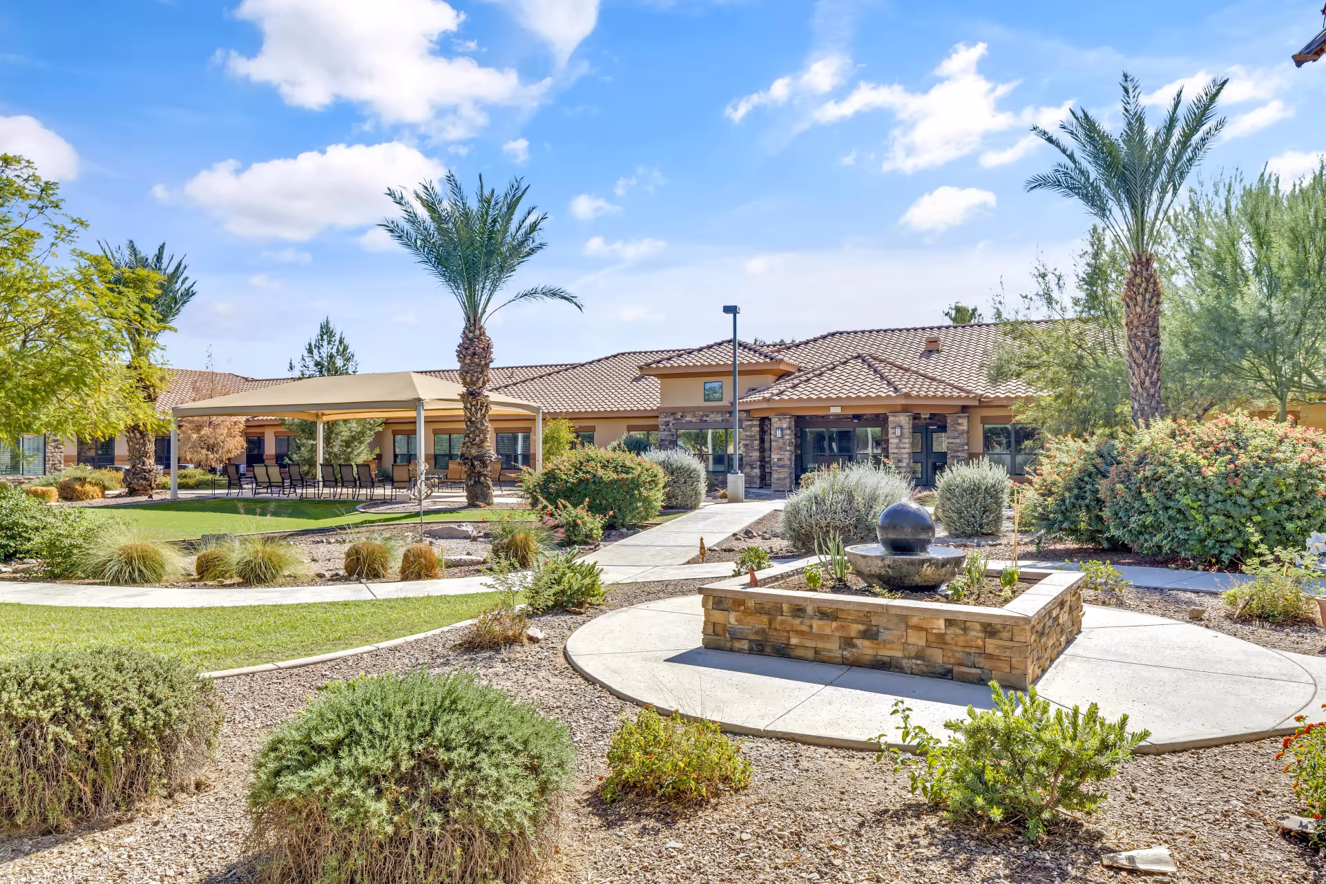 Sunny landscaped courtyard with paved walkways, a central stone fountain, palm trees, and a covered patio in front of a single-story building.