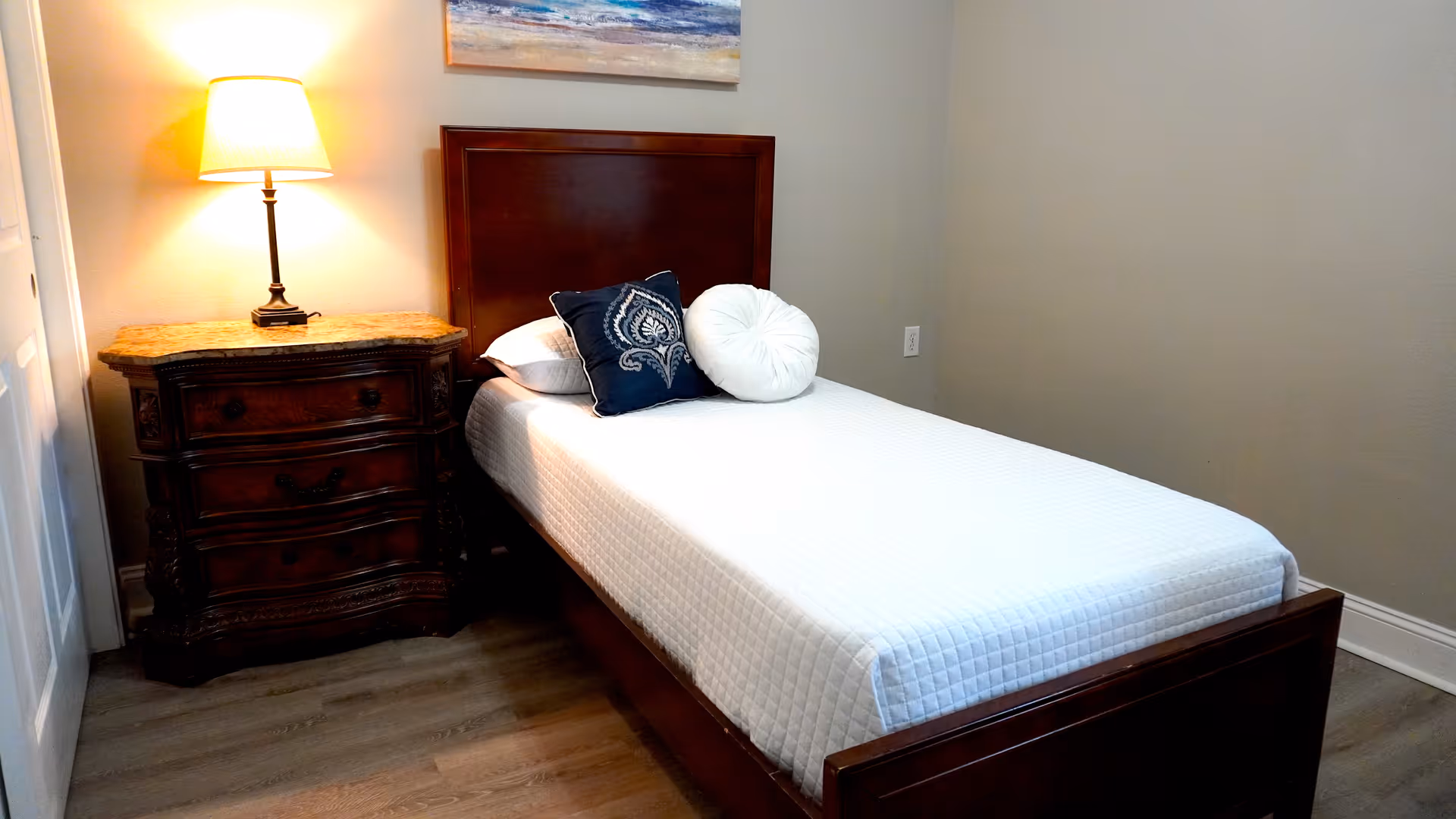 A single bed with a wooden headboard and footboard, covered with a white quilted bedspread and two decorative pillows, one navy blue with a white pattern and one round white pillow. Next to the bed is a wooden nightstand with three drawers and a marble top, holding a lit table lamp. The room has light-colored walls and wood flooring, with a painting hanging above the bed.