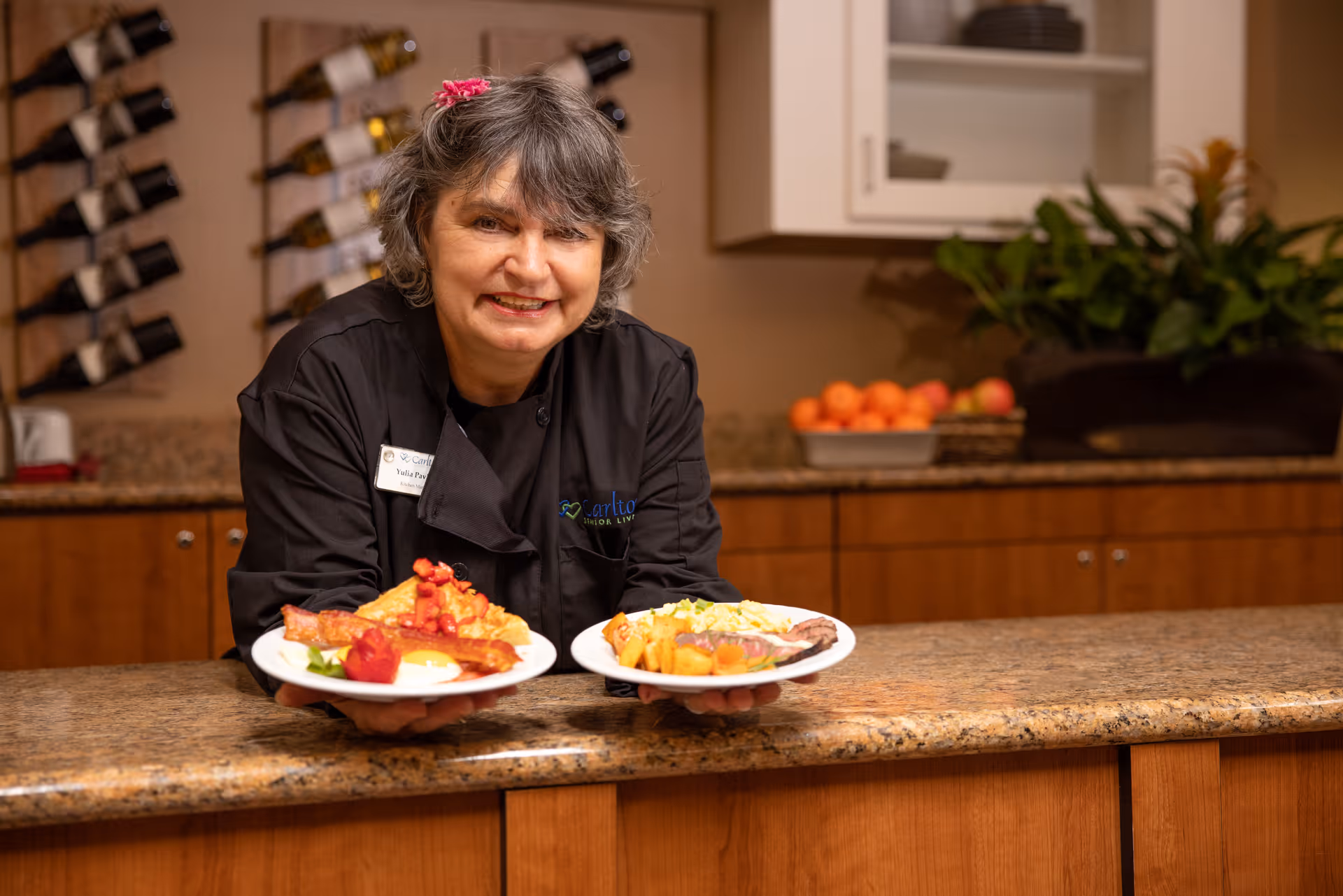 A smiling woman wearing a black chef's coat with a name tag that reads 'Yulia' is standing behind a kitchen counter holding two plates of food. The plates contain breakfast items including bacon, eggs, and fruit. Behind her, there is a wine rack on the wall, a bowl of oranges on the counter, and some plants.
