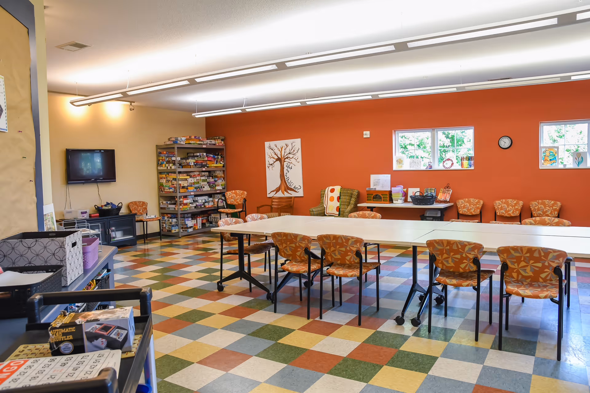 A brightly lit activity room with colorful checkered flooring, orange patterned chairs around white tables, a bookshelf filled with board games, a wall-mounted TV, and windows letting in natural light. The walls are painted orange and beige, and there is a painting of a tree on the wall.