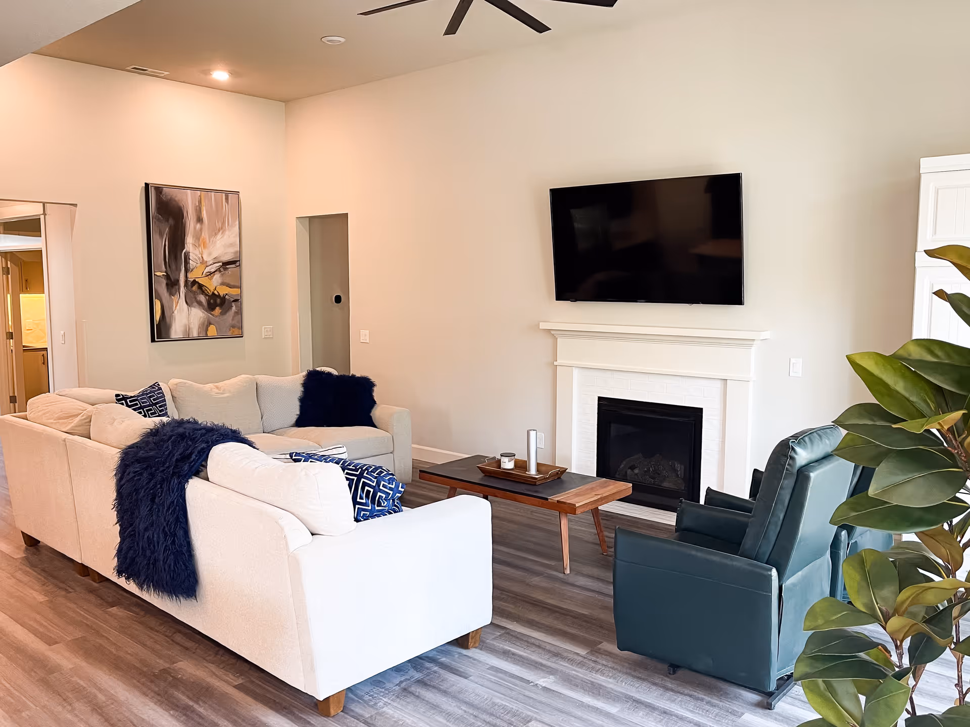A modern living room with light-colored walls and wood flooring. The room features a beige sectional sofa with dark blue throw pillows and a matching throw blanket, a wooden coffee table with decorative items, a dark green recliner chair, a white fireplace with a mounted flat-screen TV above it, a large abstract painting on one wall, and a green leafy plant in the corner.