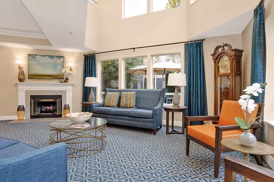 A bright and spacious living room with a high ceiling and large windows. The room features a blue sofa with striped cushions, an orange armchair, a glass coffee table, and a grandfather clock. There is a fireplace with a painting above it and two wall sconces on either side. Blue curtains frame the windows, and a patterned carpet covers the floor. Outside the windows, patio umbrellas and greenery are visible.
