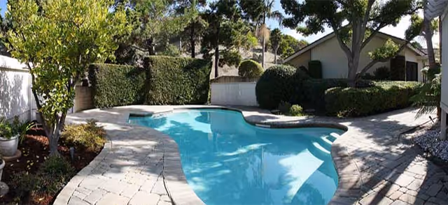 Outdoor swimming pool with a curved shape surrounded by a stone-paved patio, trees, bushes, and a small building in the background under a clear sky.