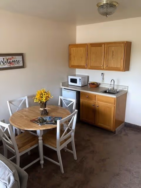 Small kitchenette area with wooden cabinets, a microwave, a mini refrigerator, and a sink. In front of the kitchenette is a round wooden table with four white chairs. A vase with yellow flowers is on the table along with some magazines. A framed 'Welcome' sign is hanging on the wall.