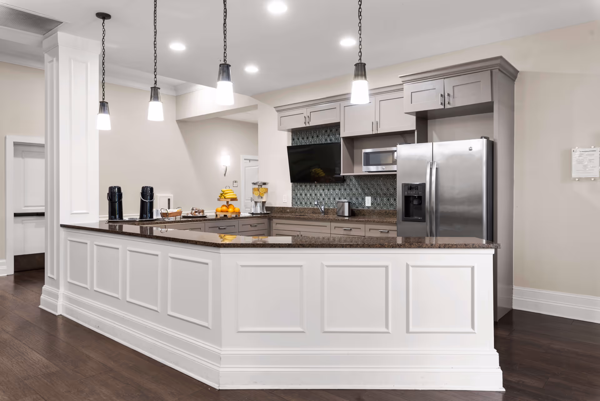 Modern kitchen area with a large white paneled island countertop, three hanging pendant lights, stainless steel refrigerator, microwave, and a wall-mounted TV. The kitchen has light gray cabinets, a dark granite countertop, and a decorative backsplash. There are coffee machines, a fruit basket, and a water dispenser on the counter.