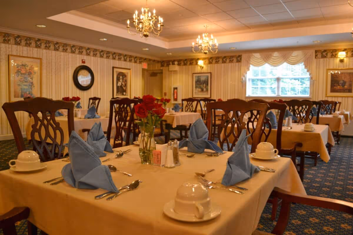 A dining room in a senior living facility with tables covered in beige tablecloths, each set with blue folded napkins, white cups and saucers, silverware, and a vase of red roses. The room has patterned carpet, wallpaper with a floral border, framed artwork on the walls, and chandeliers hanging from the ceiling. A window with white blinds and a valance lets in natural light.