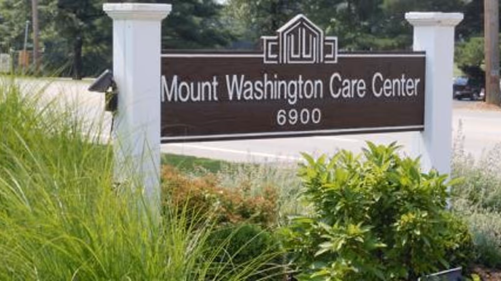 Outdoor sign for Mount Washington Care Center with the address number 6900, surrounded by green plants and grass, with a road and trees in the background.