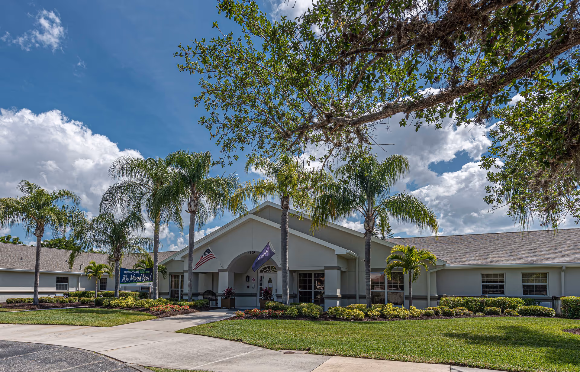 Exterior view of Arden Courts - ProMedica Memory Care Community in Sarasota, showing a single-story building with a gabled entrance, surrounded by palm trees and well-maintained landscaping under a partly cloudy blue sky.