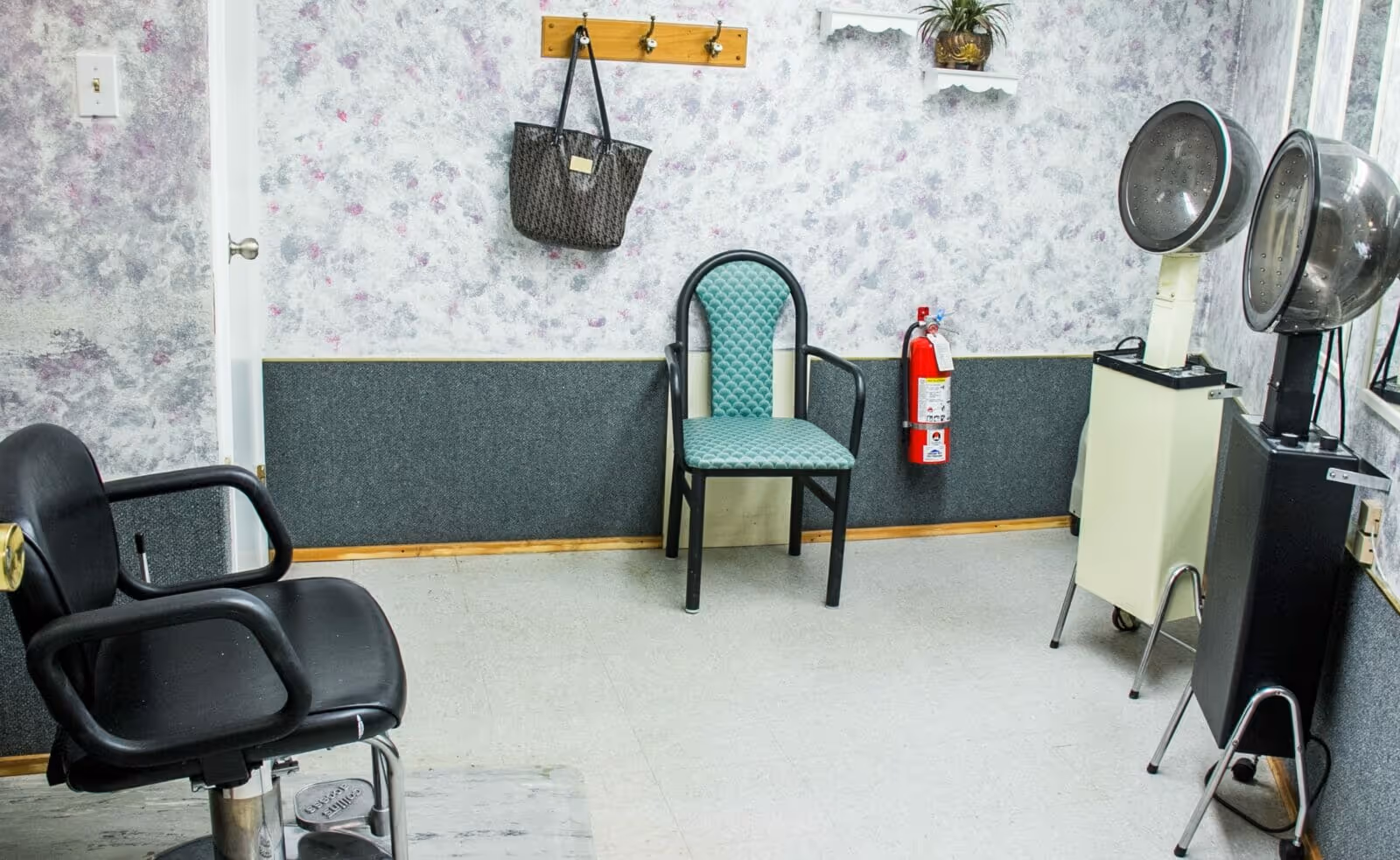 Interior of a salon or hair care room with two hair drying stations, a black salon chair, a green cushioned chair, a fire extinguisher mounted on the wall, a wall hook with a handbag hanging, and floral patterned wallpaper.