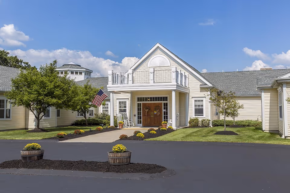 Front exterior view of a single-story senior living facility building with beige siding, a peaked roof, and a covered entrance supported by white columns. The entrance features double wooden doors with wreaths, two white rocking chairs, and an American flag mounted on the left side. The building is surrounded by well-maintained green lawns, small trees, and flower beds with yellow and orange flowers. The sky is blue with scattered clouds.