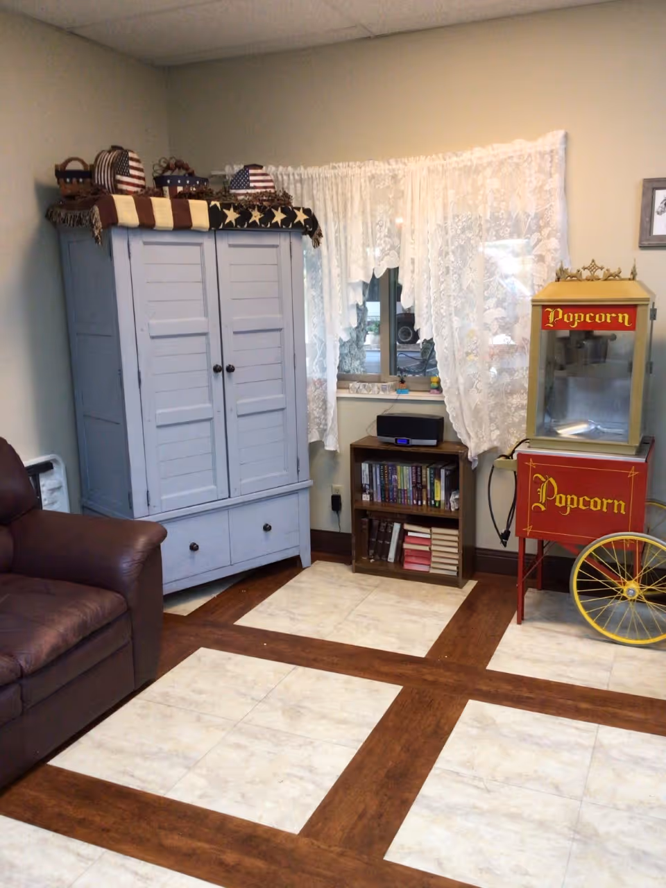 Small sitting area with a brown leather armchair, pale blue cabinet, bookshelf under a lace-curtained window, and a red vintage popcorn machine on a checkered wood-and-tile floor.