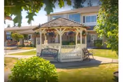 A white wooden gazebo with a shingled roof situated on a paved area surrounded by green grass and bushes. There are benches inside and outside the gazebo. In the background, there is a two-story building with multiple windows and a covered porch area. Trees and shrubs frame the scene under a clear sky.