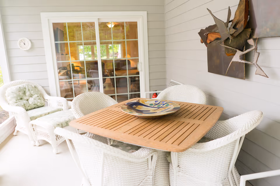 Enclosed porch with a wooden dining table surrounded by white wicker chairs and a decorative bowl, and sliding glass doors leading inside.