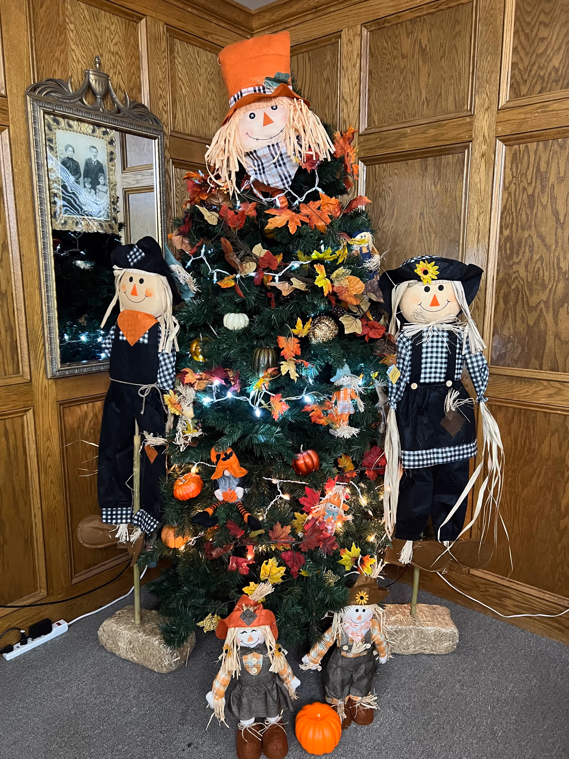 A decorated artificial tree with autumn-themed ornaments and lights, surrounded by five scarecrow dolls dressed in fall attire. The scene is set in a corner with wood-paneled walls and a framed black-and-white family photo reflected in a mirror on the left side.