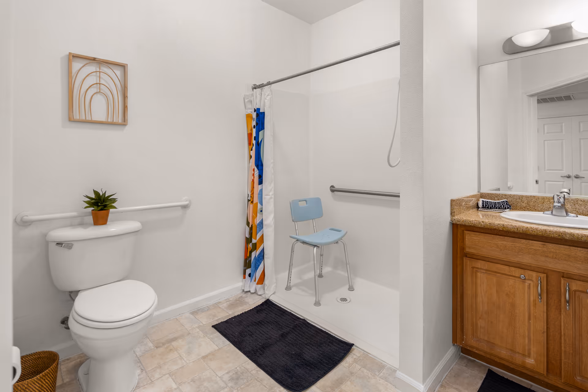 A clean and accessible bathroom featuring a white toilet with a small potted plant on top, a walk-in shower with a blue shower chair and colorful shower curtain, a wooden vanity with a granite countertop and sink, a large mirror above the sink, and a wall-mounted light fixture. The floor is tiled and there are black bath mats on the floor.