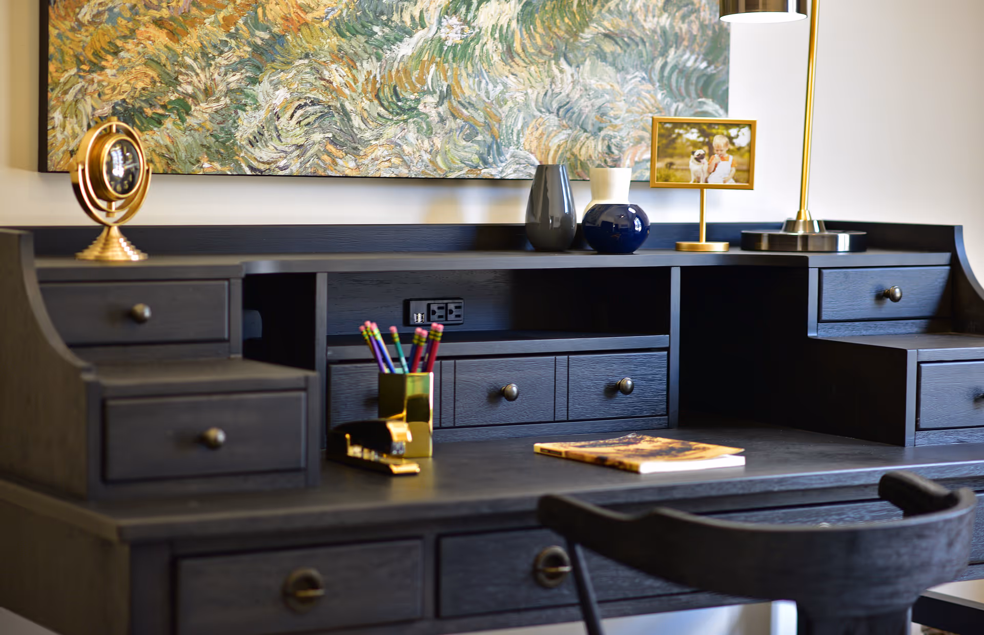 A dark wooden desk with small drawers holds a lamp, framed photo, vases, pencils, and a book beneath a textured painting.
