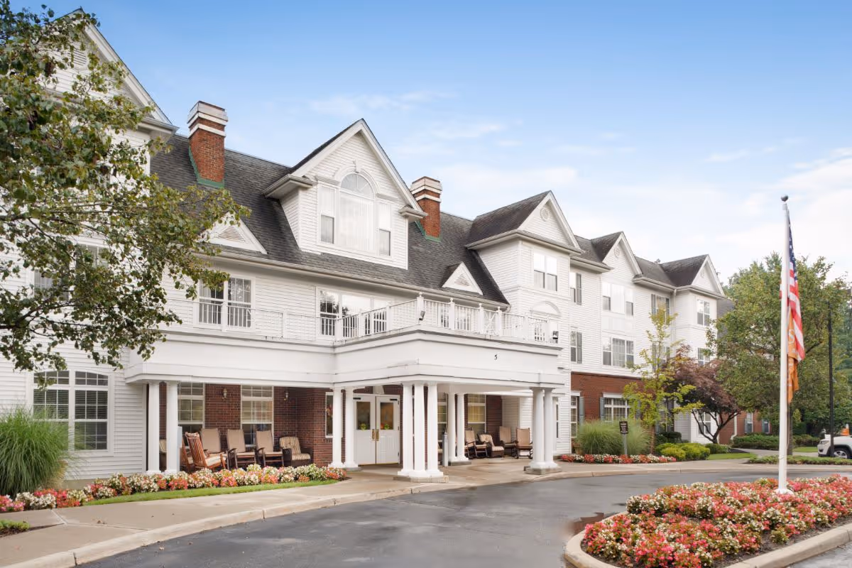 Front exterior of a large multi-story senior living facility with a covered entrance, rocking chairs, landscaped flowerbeds and an American flag.
