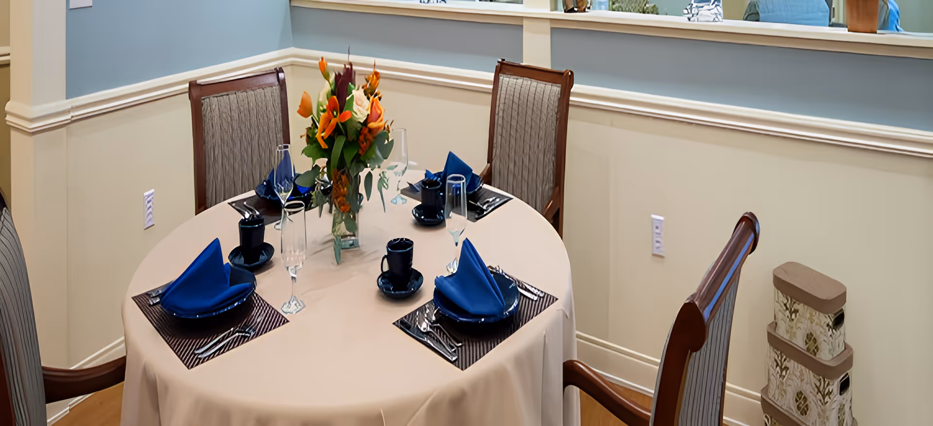 Round dining table set for four with blue napkins, dark dishes, wine glasses, and a floral centerpiece in a softly painted dining room.