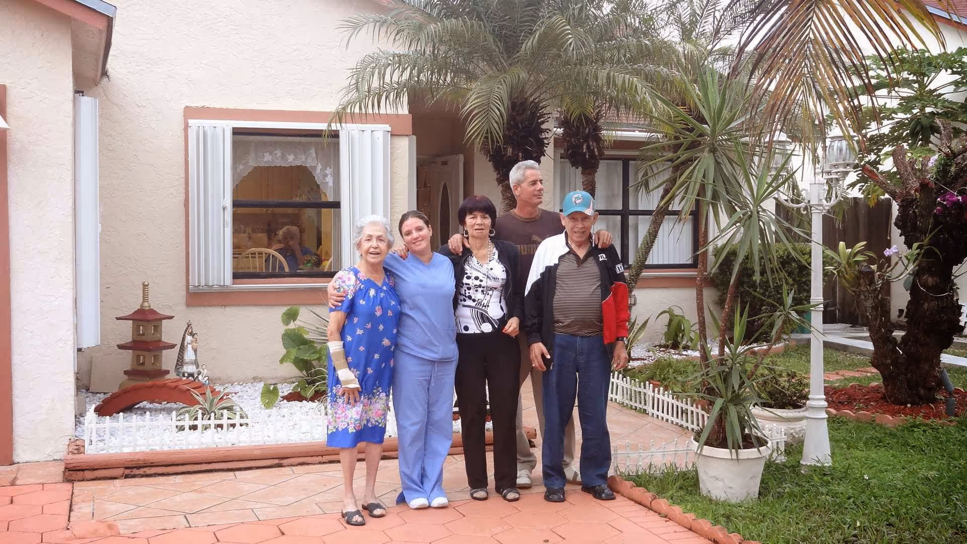 Five people standing together outside in a garden area of a residential facility. The group includes elderly individuals and a caregiver dressed in blue scrubs. Behind them is a beige building with windows, plants, and a small white fence surrounding garden beds.
