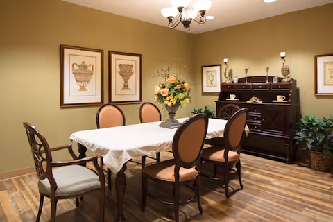 A dining room with a rectangular table covered with a white tablecloth, surrounded by six wooden chairs with cushioned seats. A floral centerpiece is on the table. The room has wooden flooring, green walls, three framed pictures on the wall, a dark wooden sideboard with decorative items, and a ceiling light fixture.