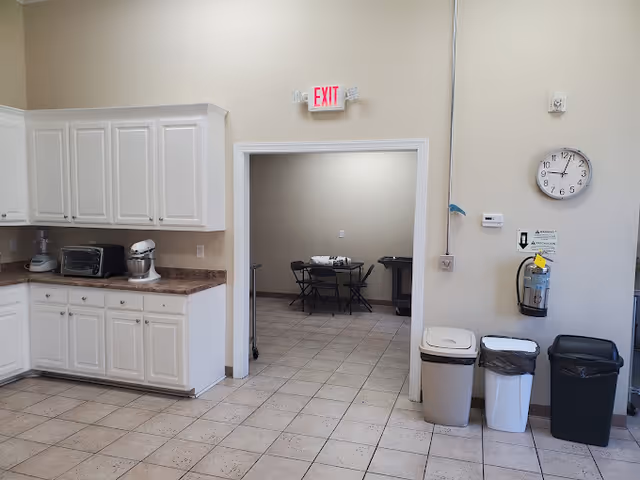 Interior view of a kitchen area with white cabinets, a toaster oven, and a stand mixer on the countertop. There is an open doorway leading to another room with a table and chairs. On the right wall, there is a clock, a fire extinguisher, and three trash bins lined up below.