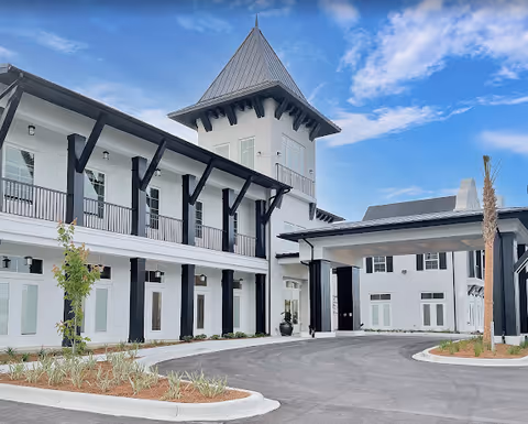 Front entrance of The Blake at Miramar Beach showing a white two-story building with a central tower, porte-cochere, and driveway under a blue sky.