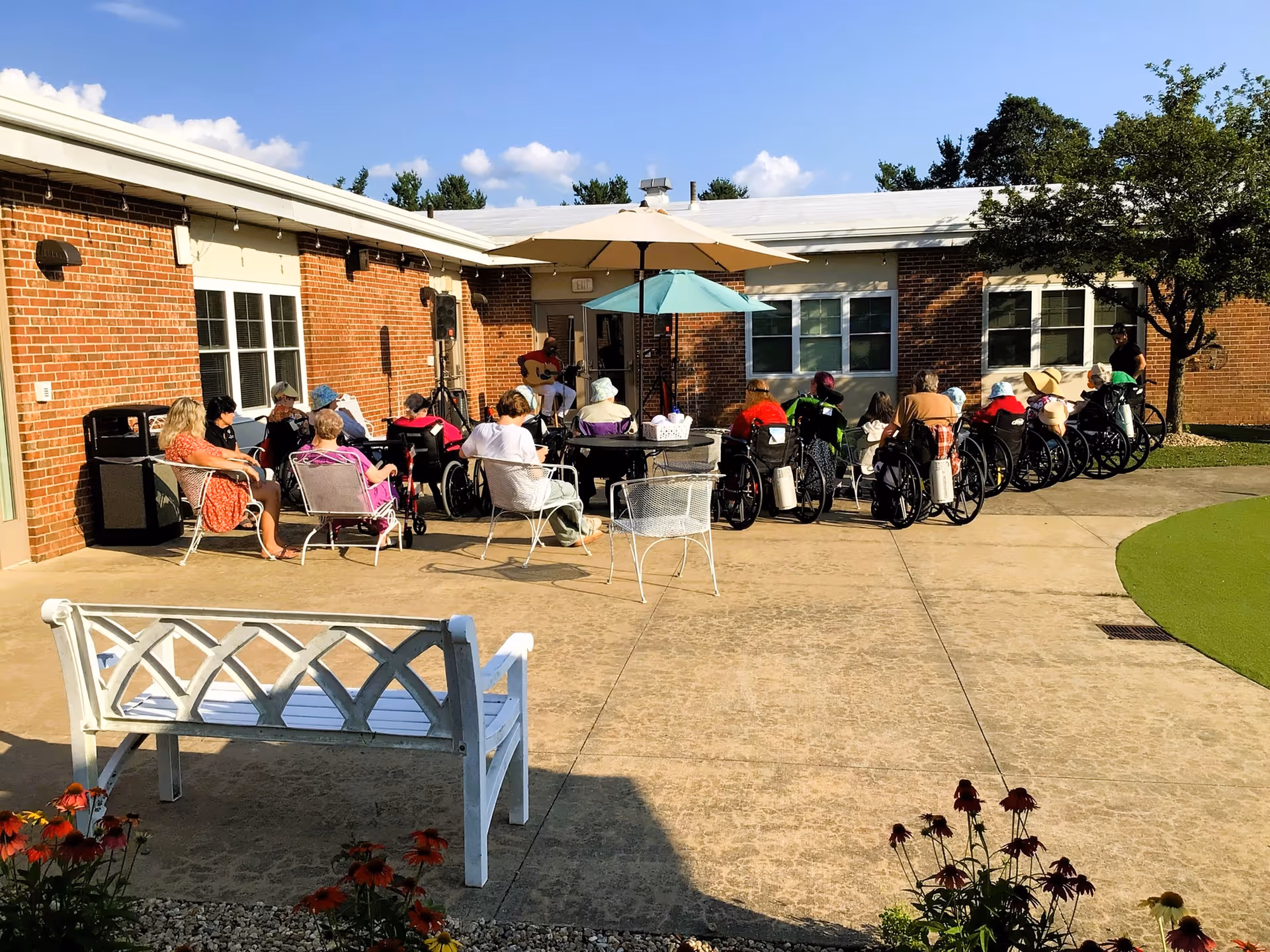Outdoor patio area at Holden Rehabilitation & Skilled Nursing Center with a group of elderly people, many in wheelchairs, seated in a semicircle facing a person playing guitar under umbrellas. The setting includes a white bench, flowers, and a brick building with windows in the background.