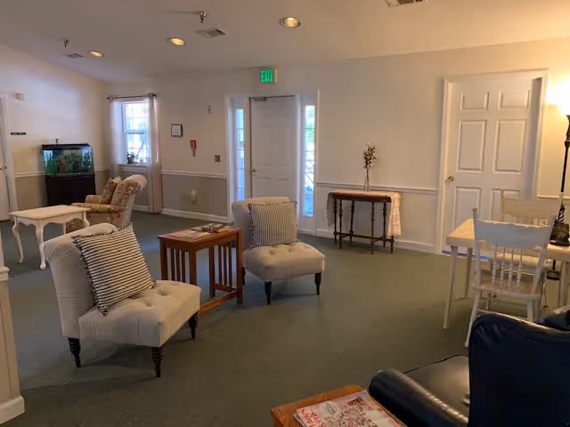 A cozy living room area with two beige cushioned chairs with striped pillows, a small wooden side table, a fish tank on a stand, a floral armchair, a white door with windows on either side, a small wooden table with a vase, and a dining table with chairs. The room has light-colored walls and green carpeted flooring.