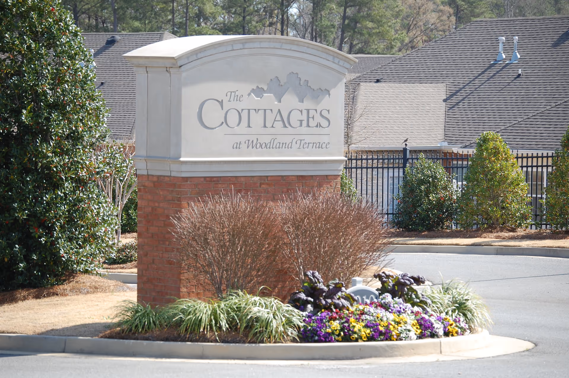 Entrance sign for The Cottages at Woodland Terrace, surrounded by landscaping with bushes and colorful flowers, with a building and trees in the background.
