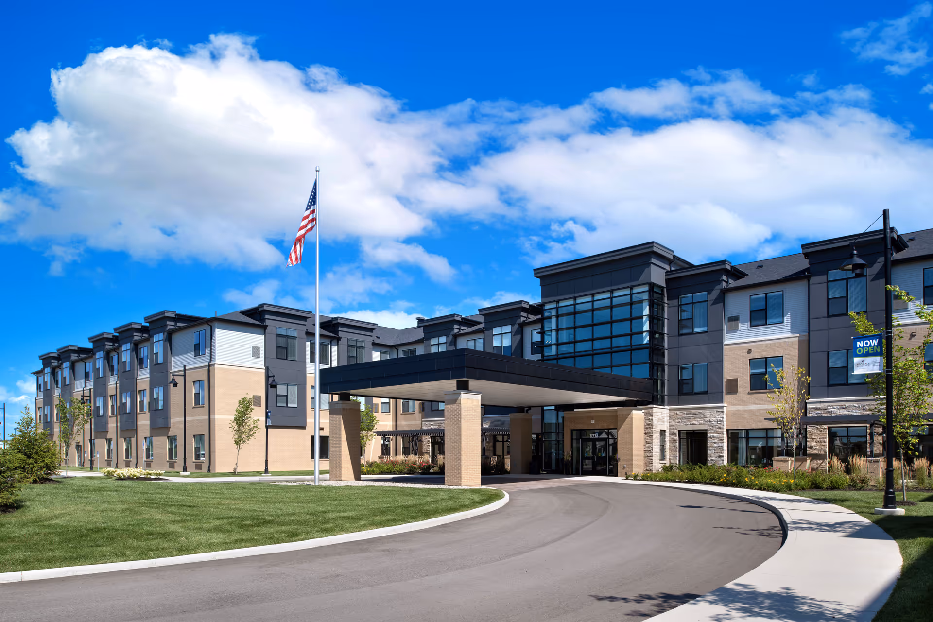 Exterior view of a modern three-story senior living facility building with a covered entrance, an American flag on a flagpole, well-maintained landscaping, and a clear blue sky with some clouds.