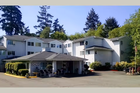 Exterior view of a multi-story senior living facility building with white siding, surrounded by trees and greenery. The entrance area has a covered porch with chairs and potted plants. The building number 10420 is visible near the entrance.