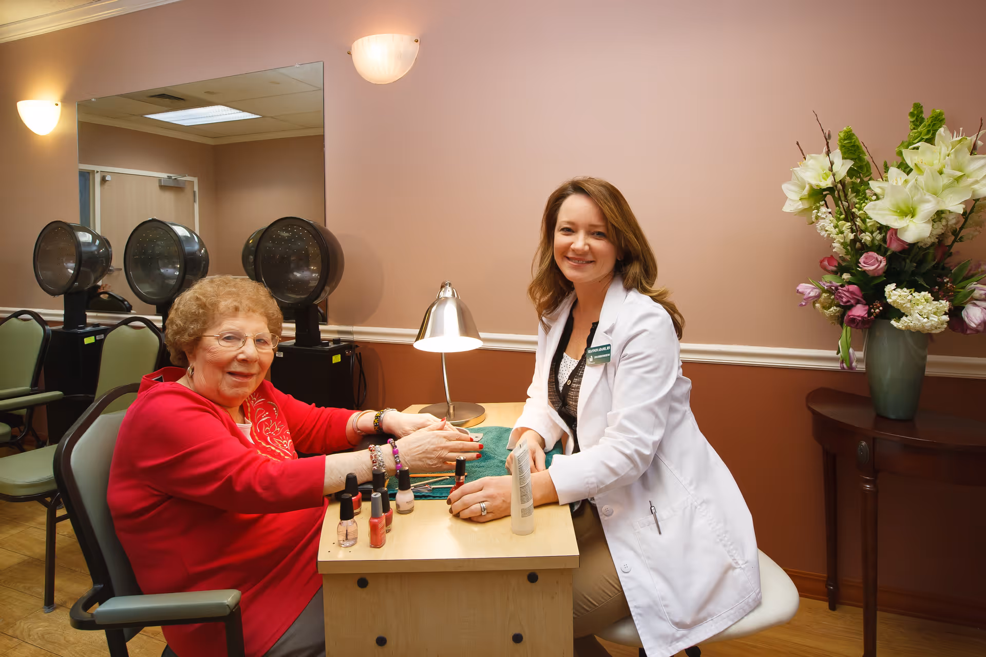 An elderly woman in a red sweater is sitting at a small table having her nails done by a smiling woman in a white coat. The setting appears to be a salon area within a senior living facility, with hair dryers and chairs visible in the background and a vase of flowers on a side table.