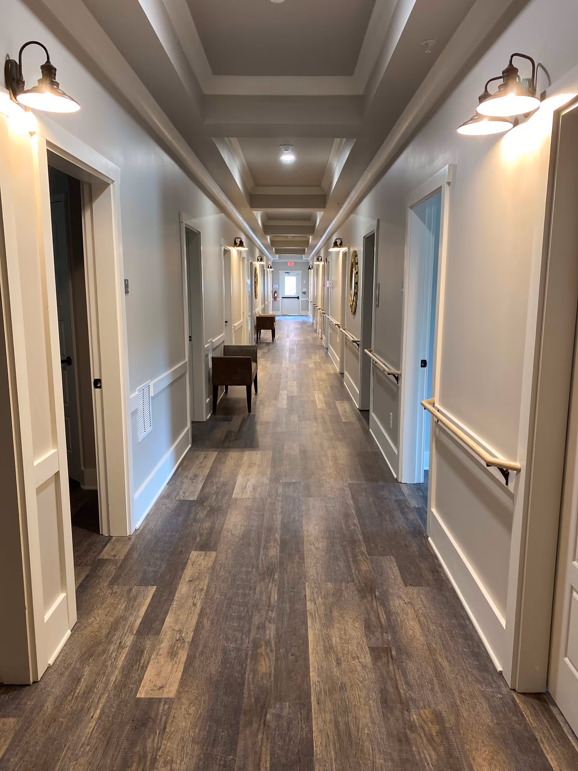 A long, well-lit hallway in a senior living facility with wood-patterned flooring, white walls, and multiple doorways on both sides. The ceiling has recessed sections with lighting, and wall-mounted lamps provide additional illumination. Handrails run along both sides of the hallway, and a few chairs are placed against the walls.