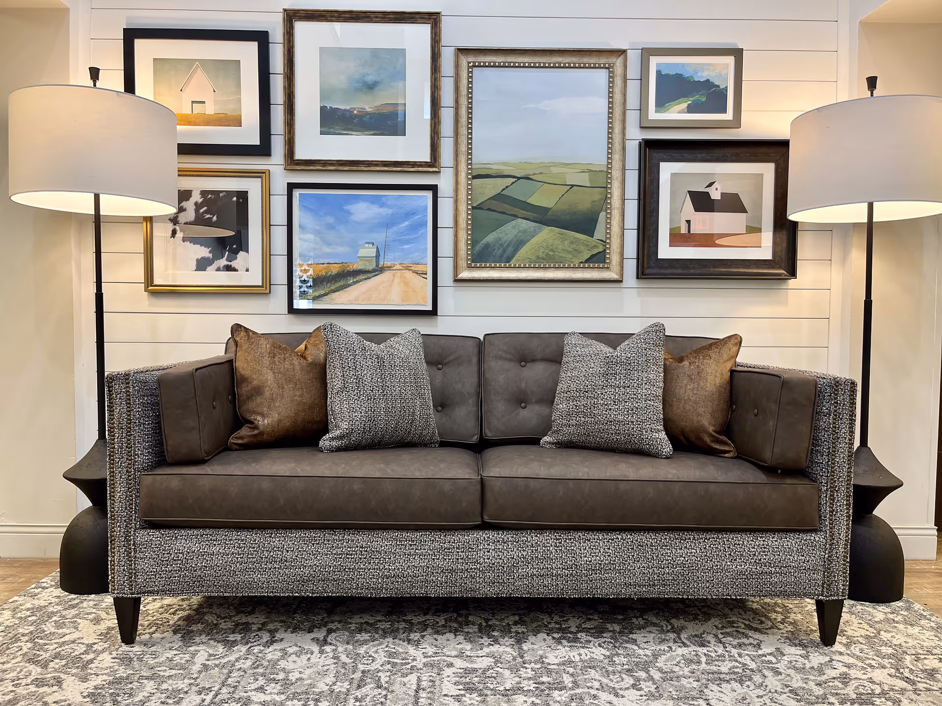 A modern living room setup featuring a gray and brown upholstered sofa with four decorative pillows. The sofa is flanked by two tall floor lamps with white lampshades. Behind the sofa is a white shiplap wall adorned with seven framed landscape and abstract art pieces arranged in a gallery style. A patterned area rug is partially visible beneath the sofa.