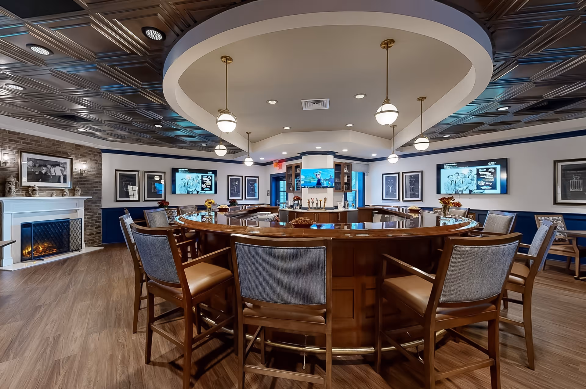 Interior view of a senior living facility lounge area featuring a large circular wooden bar with high chairs around it. The room has a decorative ceiling with hanging globe lights, a fireplace on the left wall, multiple framed pictures, and two flat-screen TVs mounted on the walls displaying black and white images. The floor is wooden, and the overall ambiance is warm and inviting.