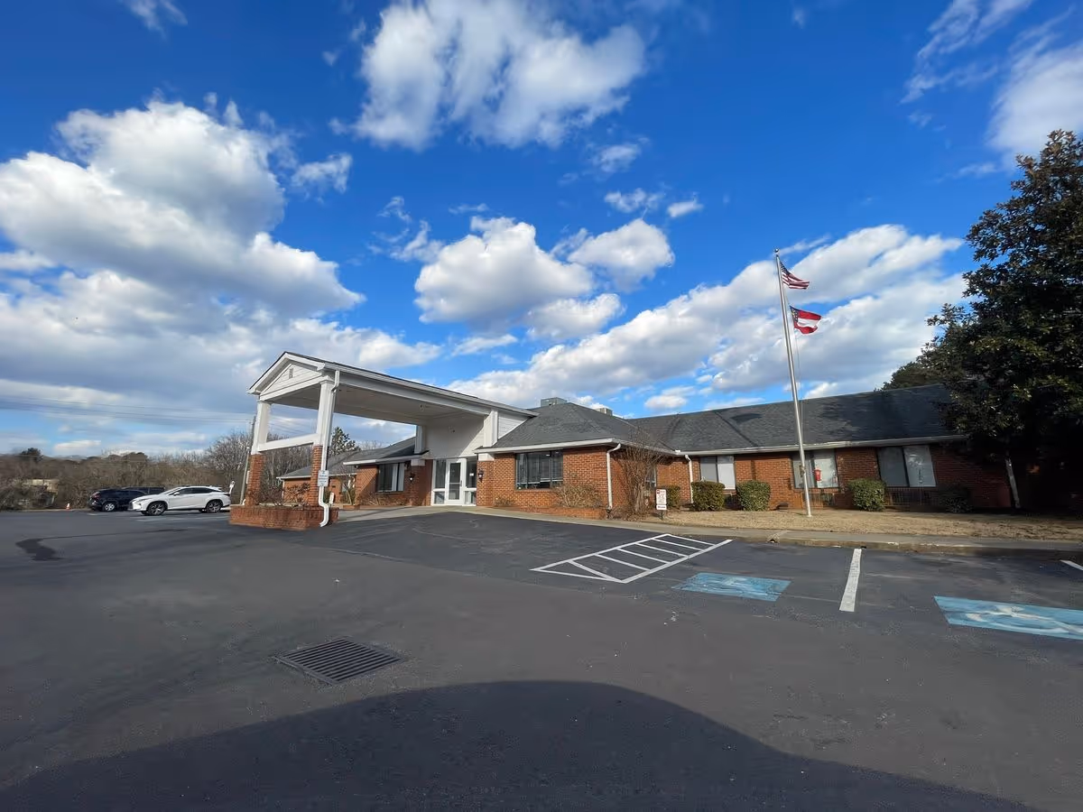 Front exterior of a single-story brick nursing facility with a covered entrance, flagpoles, and a parking lot under a blue sky.
