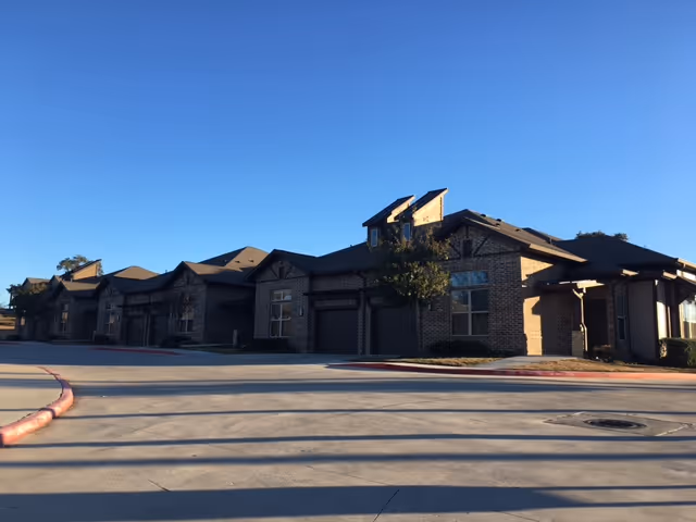 Exterior view of a single-story brick building with multiple garage doors and windows under a clear blue sky, with a concrete driveway and a small tree in front.