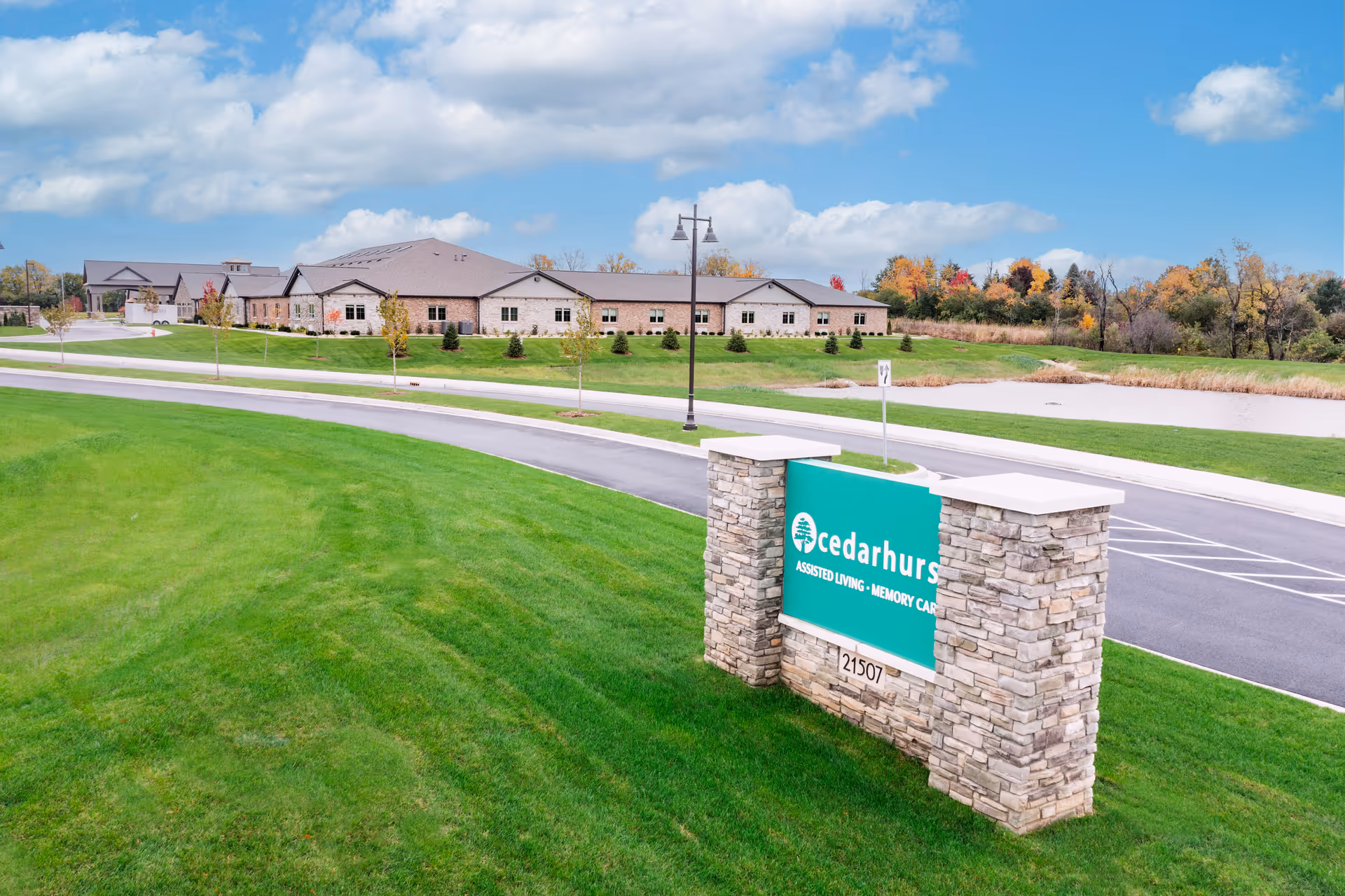 Exterior view of Cedarhurst Senior Living of Frankfort facility with a large green lawn, a stone sign displaying the facility name, a paved road, and a building in the background under a partly cloudy blue sky.
