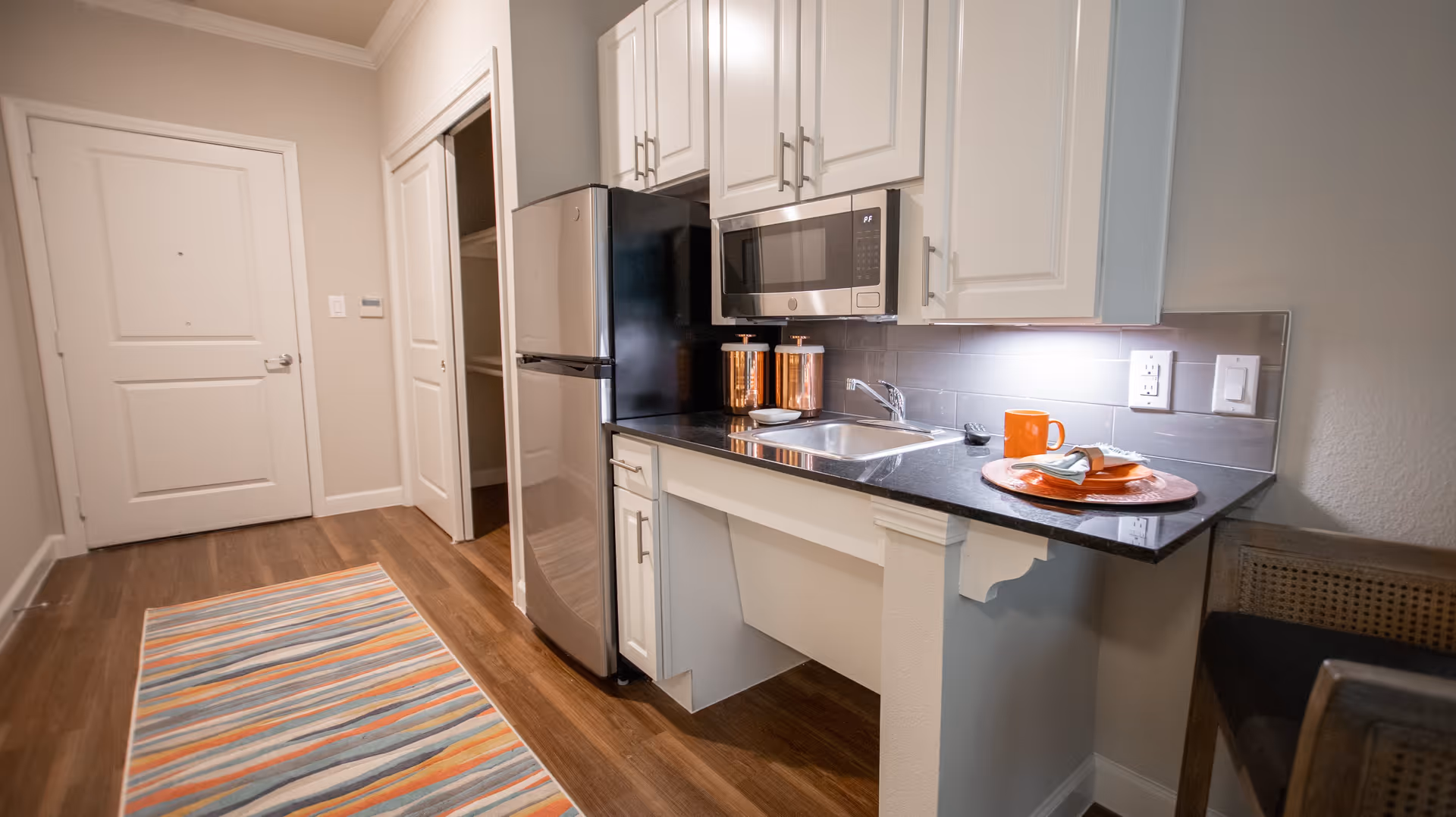 A small kitchen area with white cabinets, a stainless steel refrigerator, a microwave, and a sink. There are two copper canisters on the counter and an orange cup and plate with a napkin on a placemat. The floor is wooden with a colorful striped rug, and there is a door and a closet in the background.