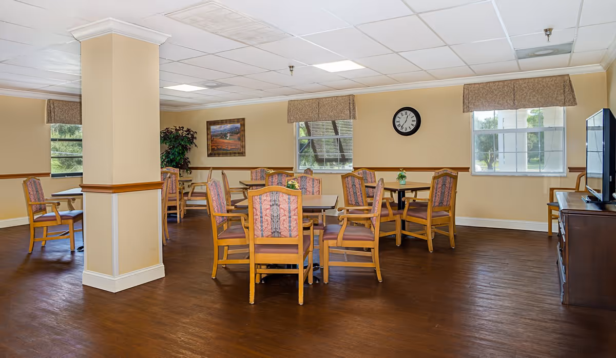 A senior living facility common area with several wooden tables and chairs arranged for seating. The room has beige walls, a dark wood floor, two windows with valances, a wall clock, a framed painting, a potted plant, and a television on a wooden stand.