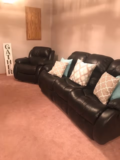 A cozy living room with a black leather sofa and matching armchair, both adorned with patterned throw pillows. The carpet is a soft pink color, and there is a wooden wall cabinet and a decorative sign that reads 'GATHER' leaning against the wall.