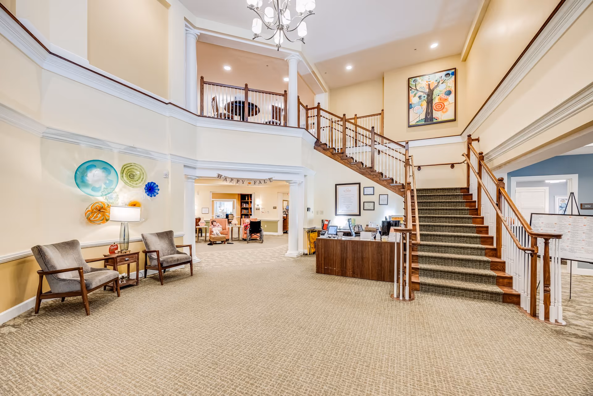 Spacious and well-lit senior living facility lobby with a carpeted floor, two gray armchairs with a small wooden table and lamp between them on the left, a wooden reception desk on the right, and a staircase with wooden handrails leading to an upper floor. The walls are decorated with colorful glass art and a framed painting, and a 'Happy Fall' banner hangs in the background above an open doorway leading to another room with seating and people.