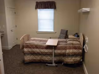 Single bedroom in a senior living room with a bed, chair, window, and small table.