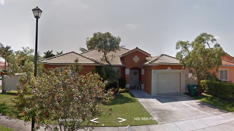 Single-story house with a tiled roof and a garage, surrounded by trees and bushes, with a street lamp on the left side and a concrete driveway leading to the garage.