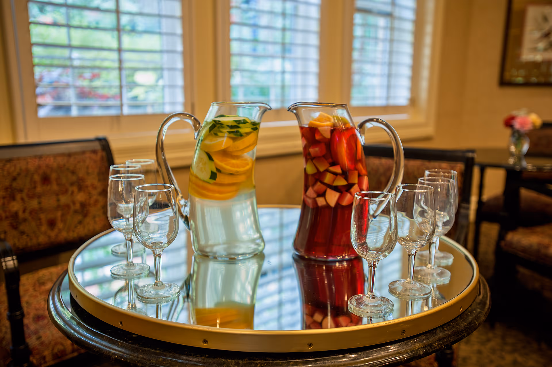 Two pitchers of fruit-infused drinks and several empty glasses arranged on a round mirrored table in a lounge area.