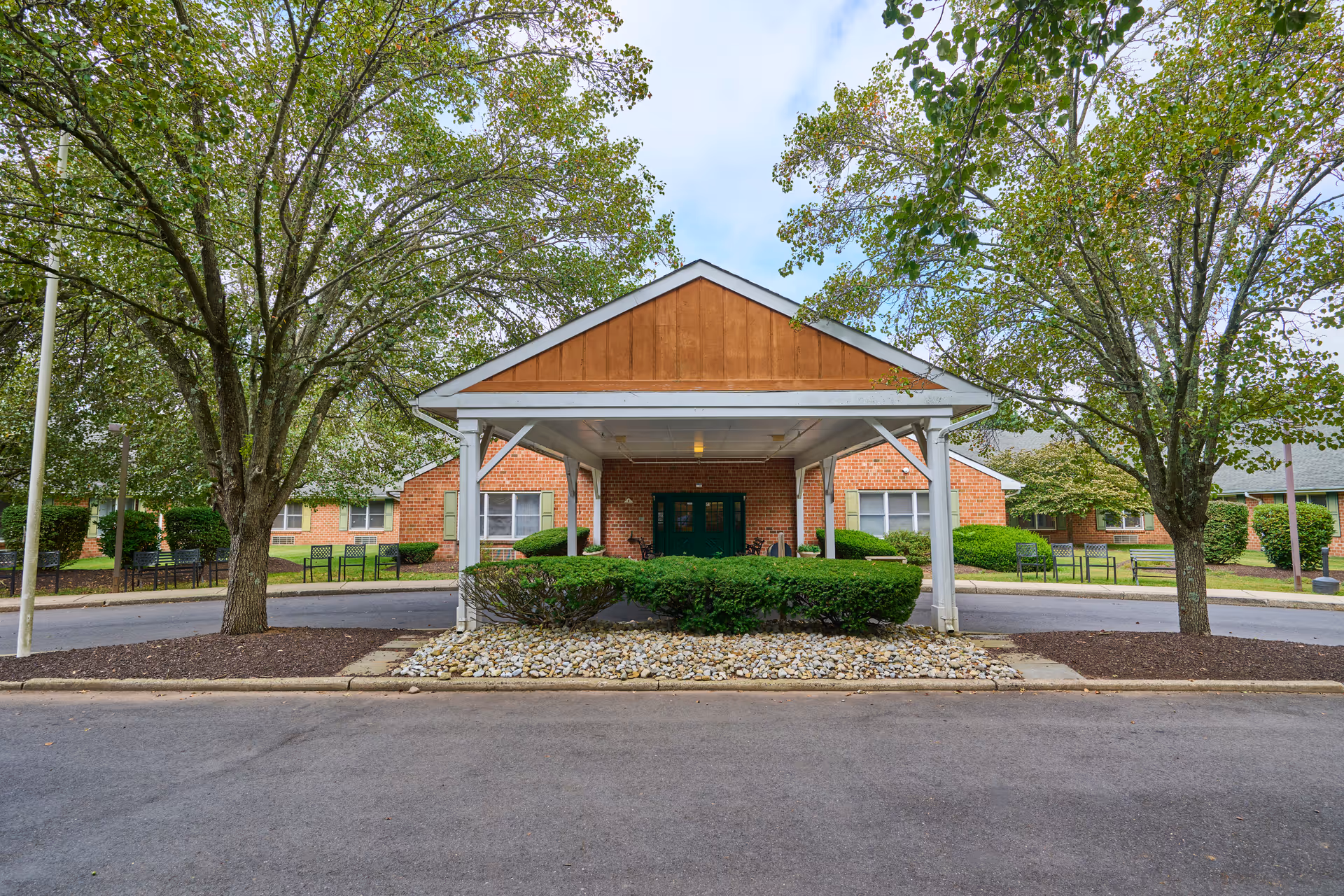 Front entrance of a brick senior care building with a covered portico, shrubs, trees, and a circular drive.