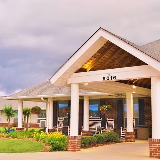 Entrance of a senior living facility with a covered porch supported by white columns and brick bases. The porch has several white rocking chairs and hanging flower baskets. The building has a gray shingled roof and brick walls, with the number 6016 displayed on the front beam of the porch roof. There is a landscaped garden with green shrubs and yellow flowers in front of the porch.