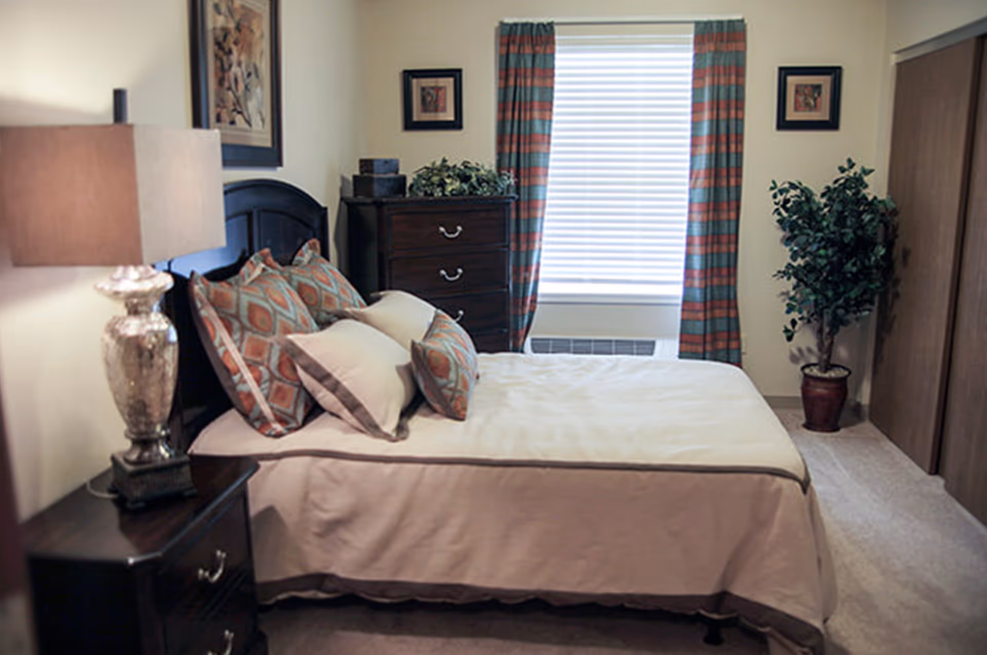 A neatly made bed with patterned pillows and a beige bedspread in a bedroom. There is a bedside table with a decorative lamp on the left, a dark wooden dresser with plants and boxes on top against the far wall, a window with striped curtains, framed artwork on the walls, and a potted plant near a wooden closet door on the right.