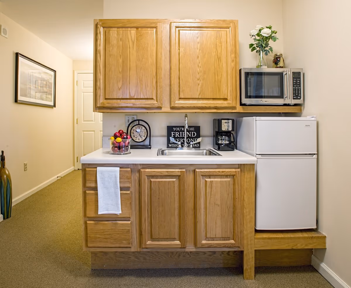 Small kitchenette with wooden cabinets, a white countertop with a sink, a glass bowl of apples, a clock, a coffee maker, a small white refrigerator, and a microwave. A white towel hangs on the cabinet handle, and a vase with white flowers is on top of the microwave. A framed picture is visible on the wall in the hallway.