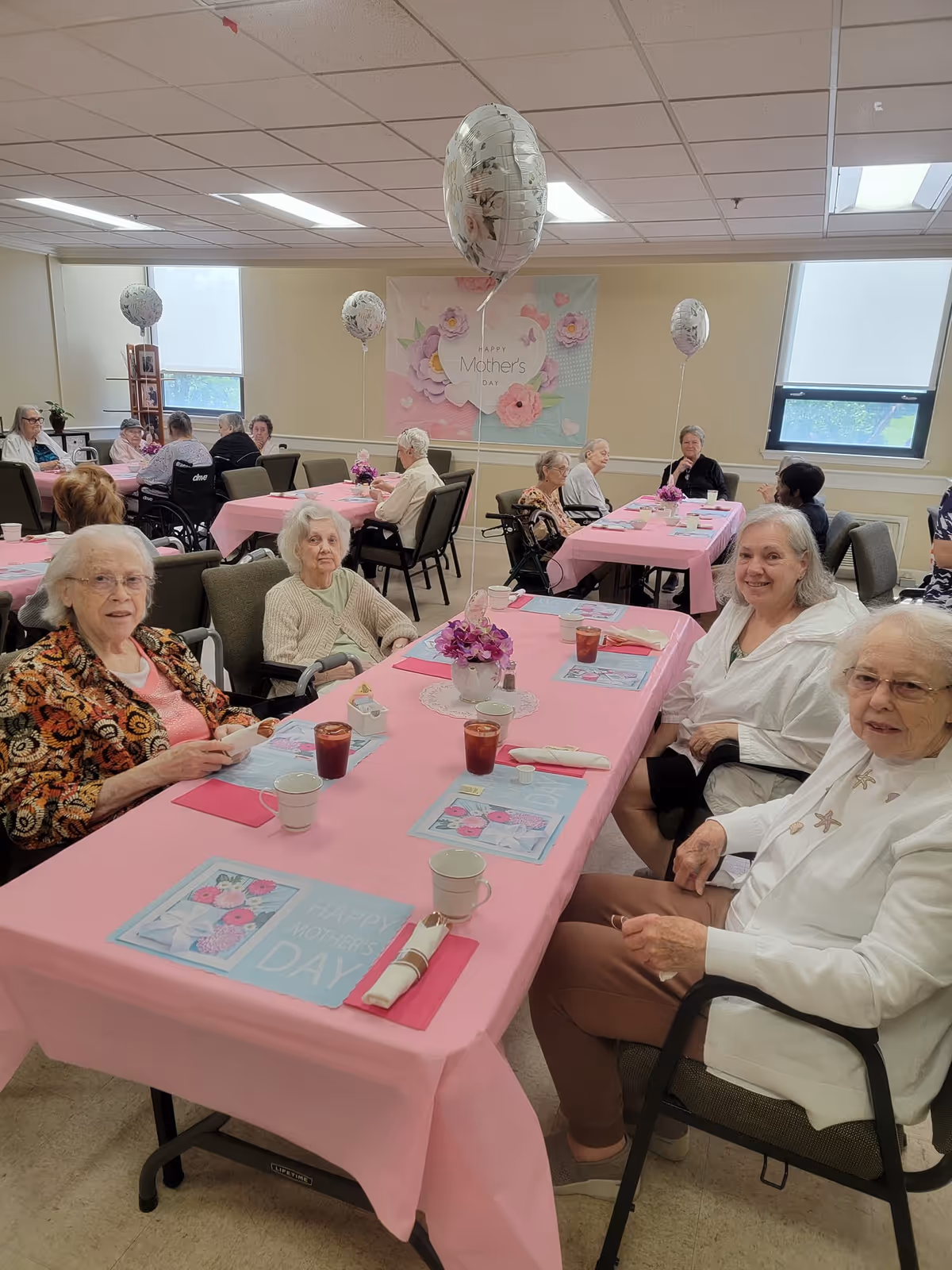 Elderly residents seated at pink-decorated tables in a common room celebrating Mother's Day with balloons and floral centerpieces.